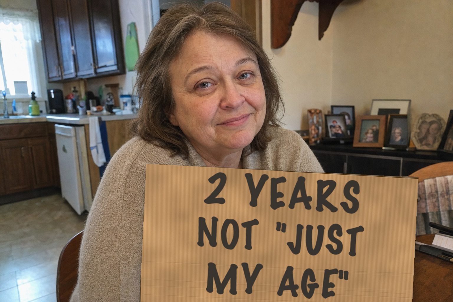 A woman at her kitchen table holding a handwritten cardboard sign reading two years, not just my age